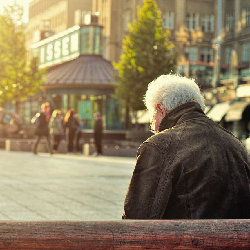 An elderly person in a leather jacket sits on a bench in public and looks down at their lap