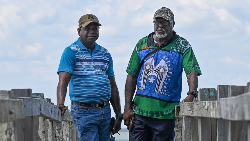 Two Torres Strait Island men posing for a photo on a boardwalk with the ocean in the background.