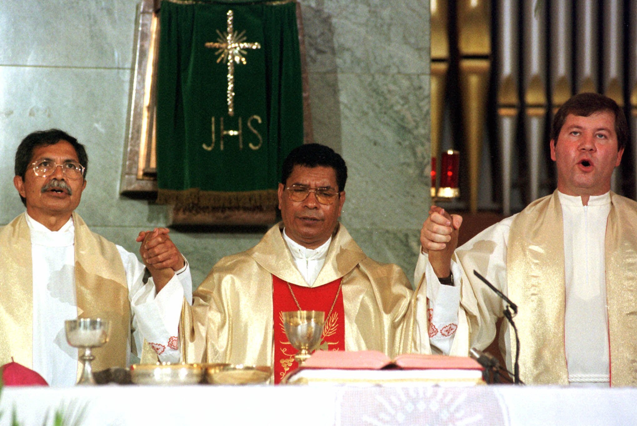 bishop in red and gold robes holds hands with two priests in front of a bible and gold goblet