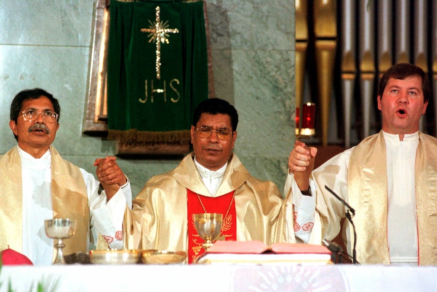 bishop in red and gold robes holds hands with two priests in front of a bible and gold goblet