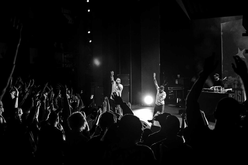 Black and white photo of rappers Briggs and Nooky waving to audience with raised arms on stage at Sydney Opera House.