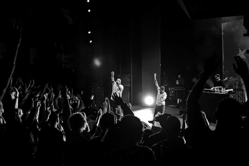 Black and white photo of rappers Briggs and Nooky waving to audience with raised arms on stage at Sydney Opera House.