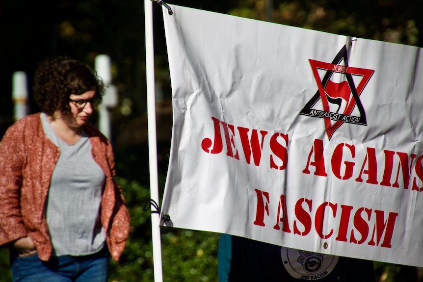 A photo of Jordy Silversteen with her hands in her pocket, walking next to a sign that reads "Jews against fascism". 