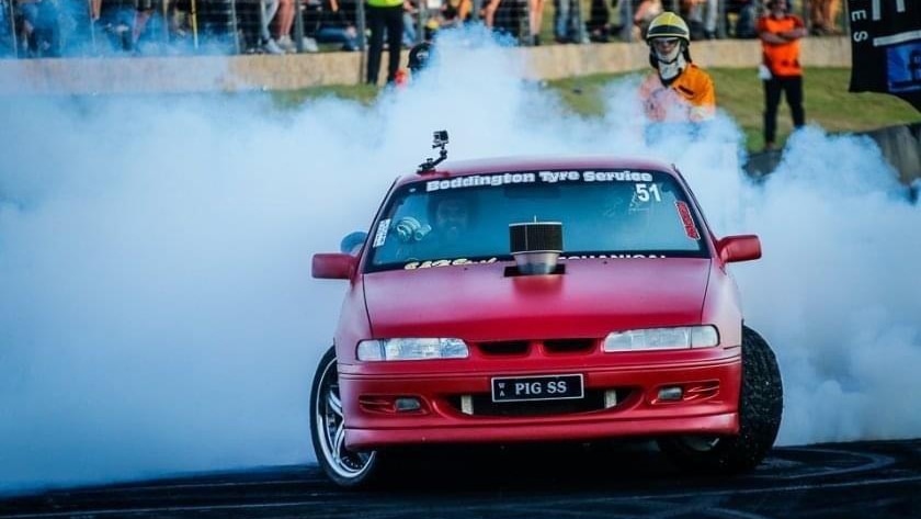 A red car does a large, smokey skid on bitumen in front of a crowd