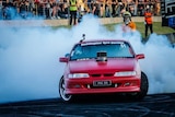 A red car does a large, smokey skid on bitumen in front of a crowd