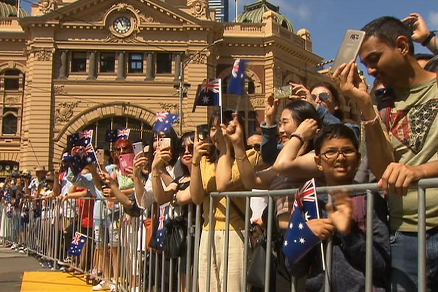 A crowd of people waving small Australian flags line the stret in front of Flinders St Station.