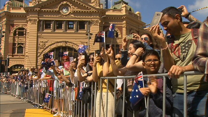 A crowd of people waving small Australian flags line the stret in front of Flinders St Station.