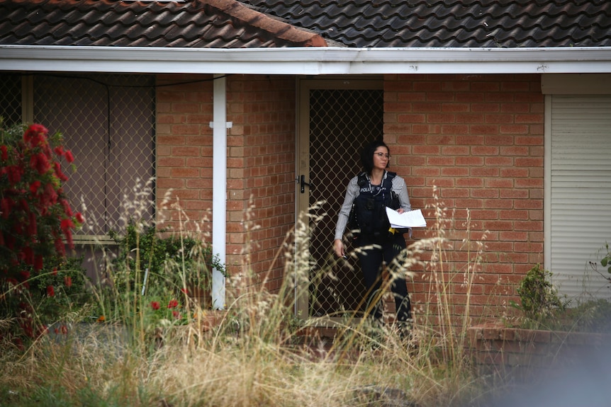 A female detective walks out of a house carrying papers with overgrown grass in the front garden.