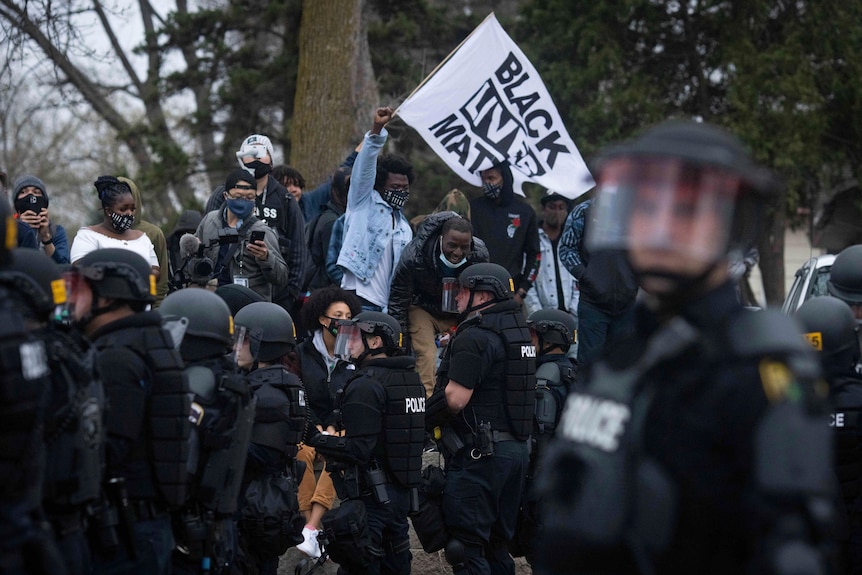 A protester raises his right fist as he stands with a crowd above a group of armed riot police.