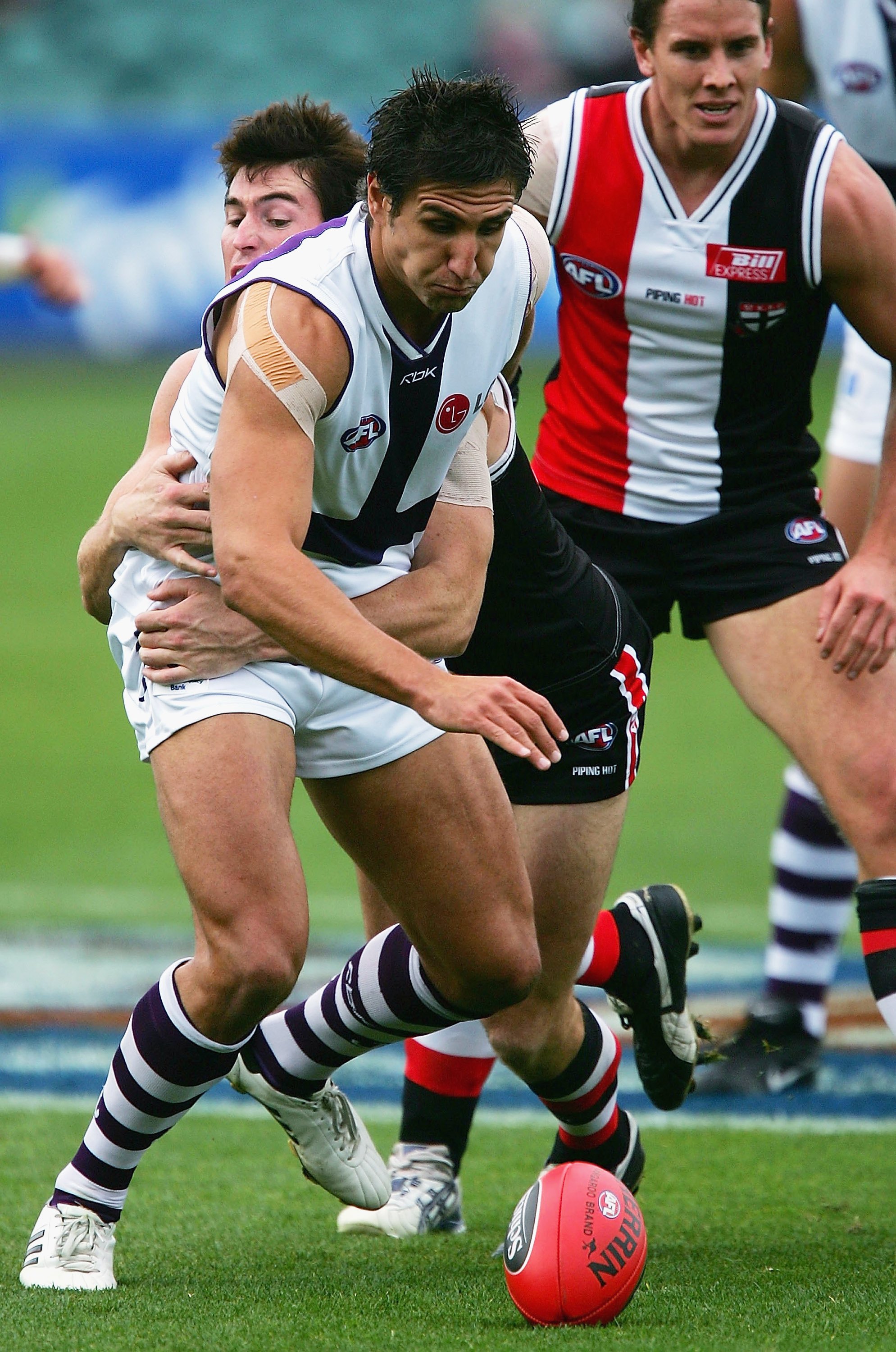 A Fremantle AFL player grimaces as he reaches for the ball while being grabbed from behind by a St Kilda defender.