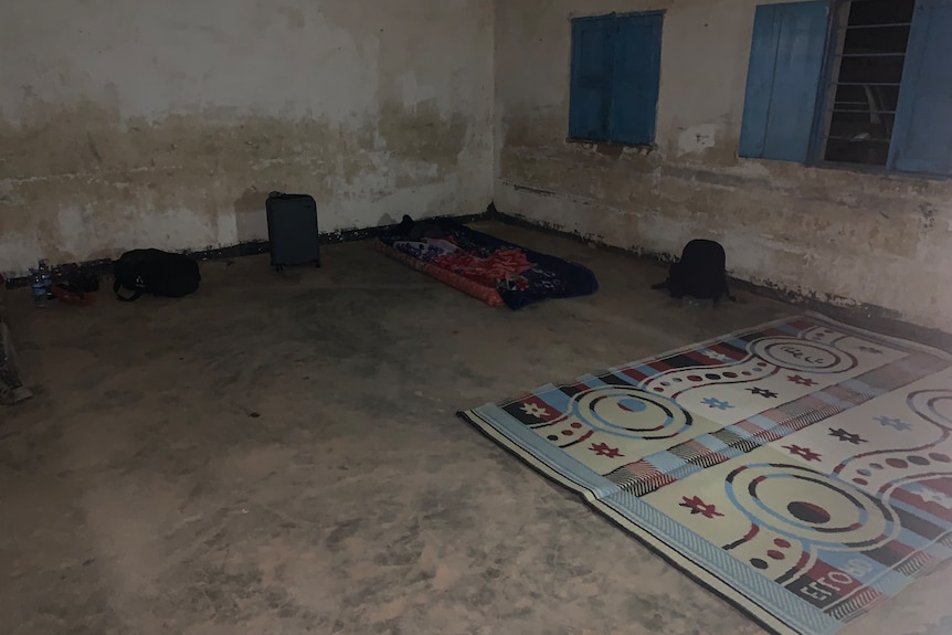 Mattress on the bare floor of a room with dirty white walls and blue window shutters.