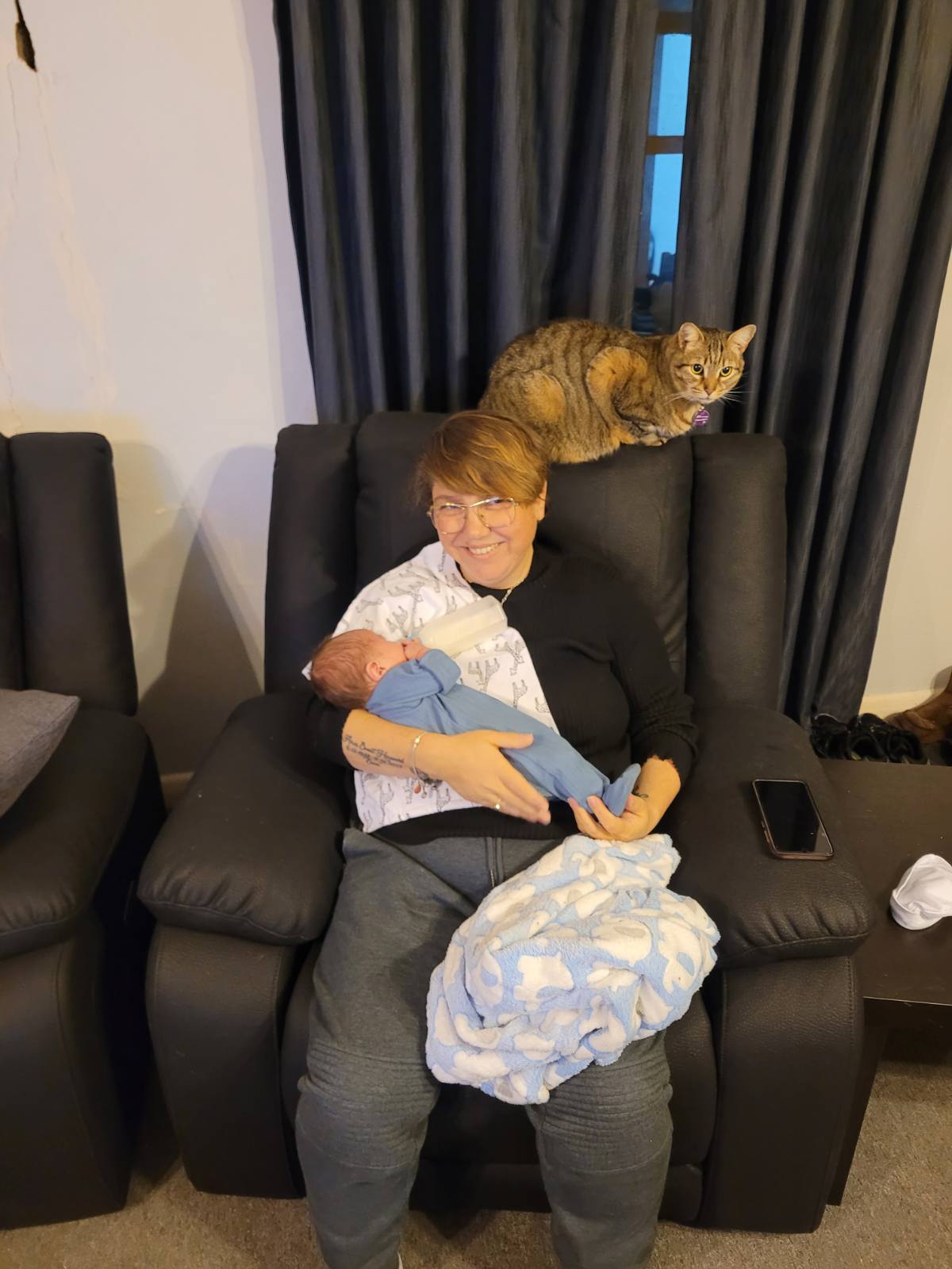 A woman with short brown hair , gold glasses, sits on couch with small baby drinking milk. A cat sits on couch behind her