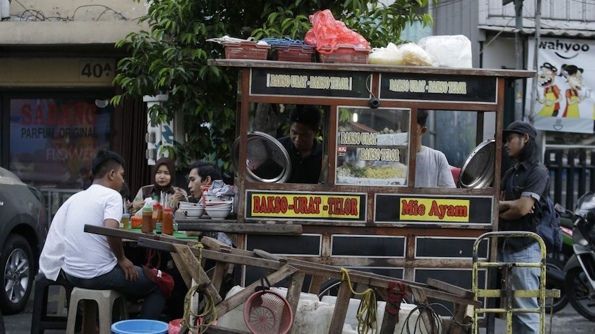 A street-vendor cart featuring signs that read 'Bakso' and 'mie ayam' surrounded by customers eating at plastic stools.