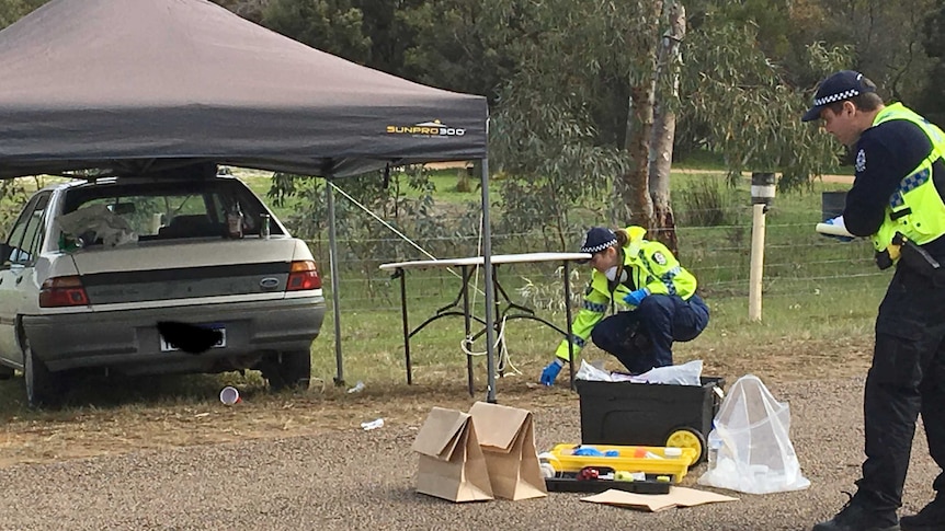 Two police officers with paper bags of evidence near a crashed car that has a tent over it.