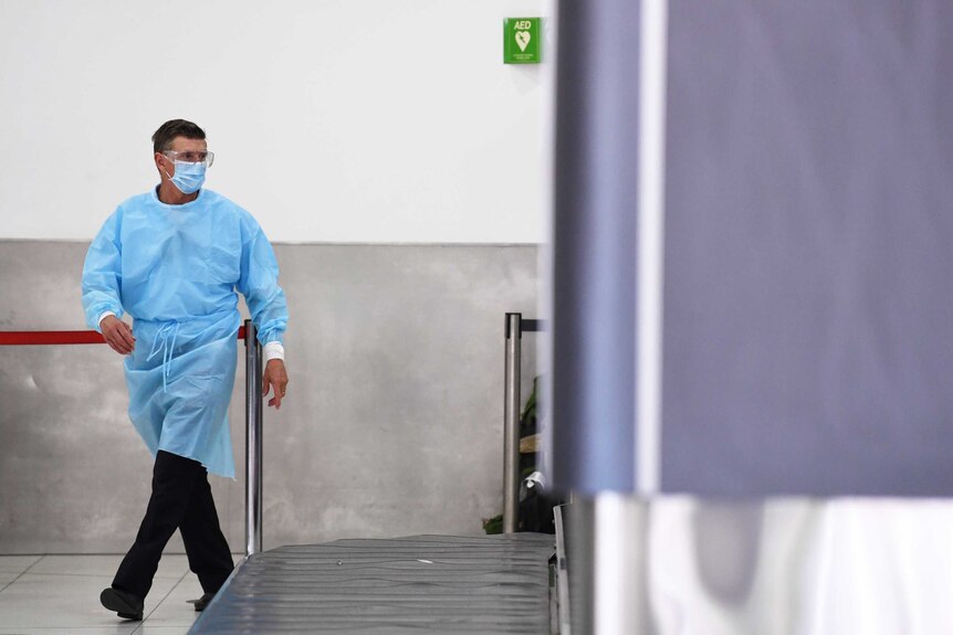A man in blue PPE walks past a baggage carousel at an airport.