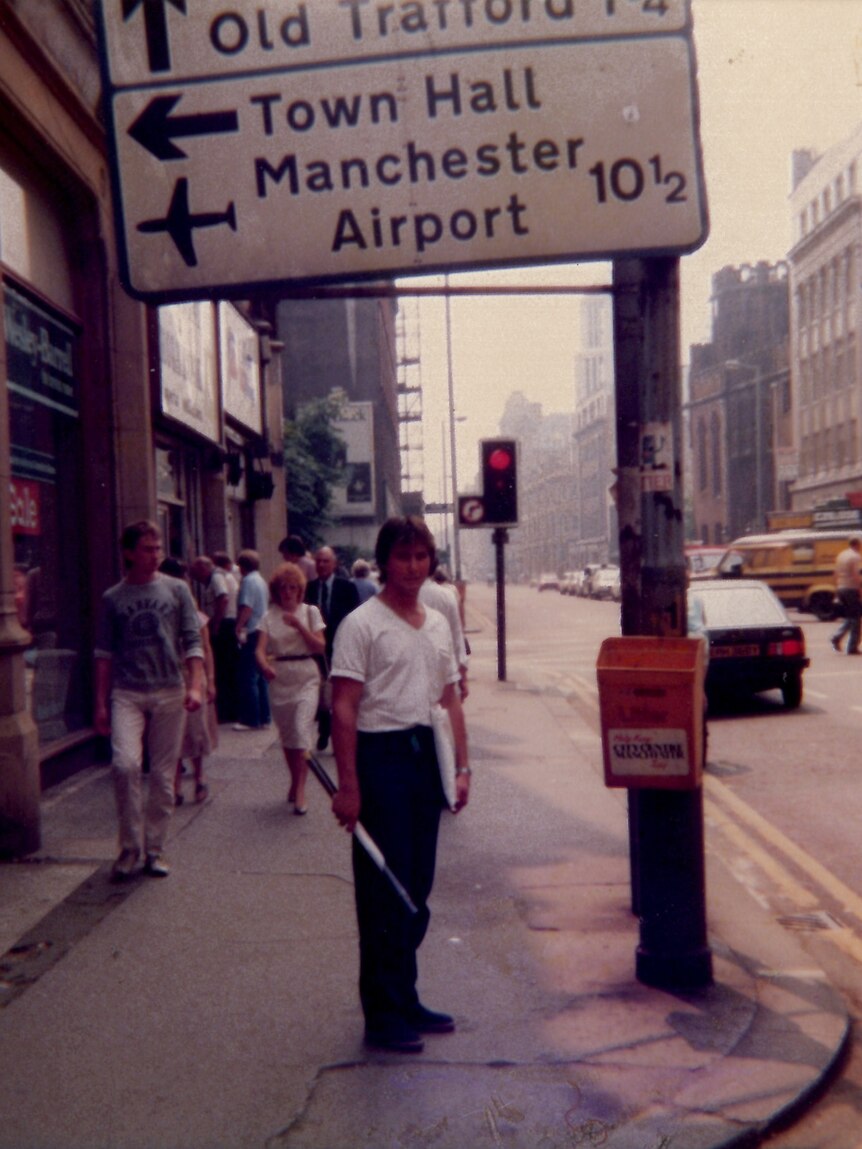 A young man stands under a sign pointing to Manchester Airport.