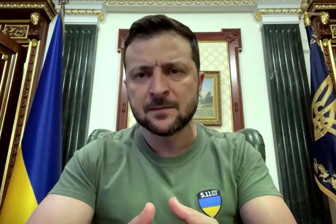 Volodymyr Zelenskyy in a green military shirt at his desk with flags in the background