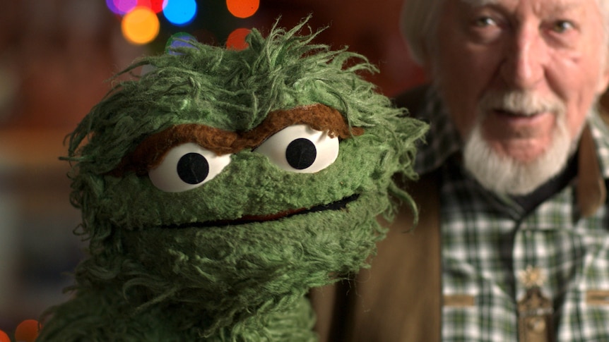 A scruffy green puppet monster with big eyes and a brown mono-brow sits beside a white-bearded man in checkered shirt.
