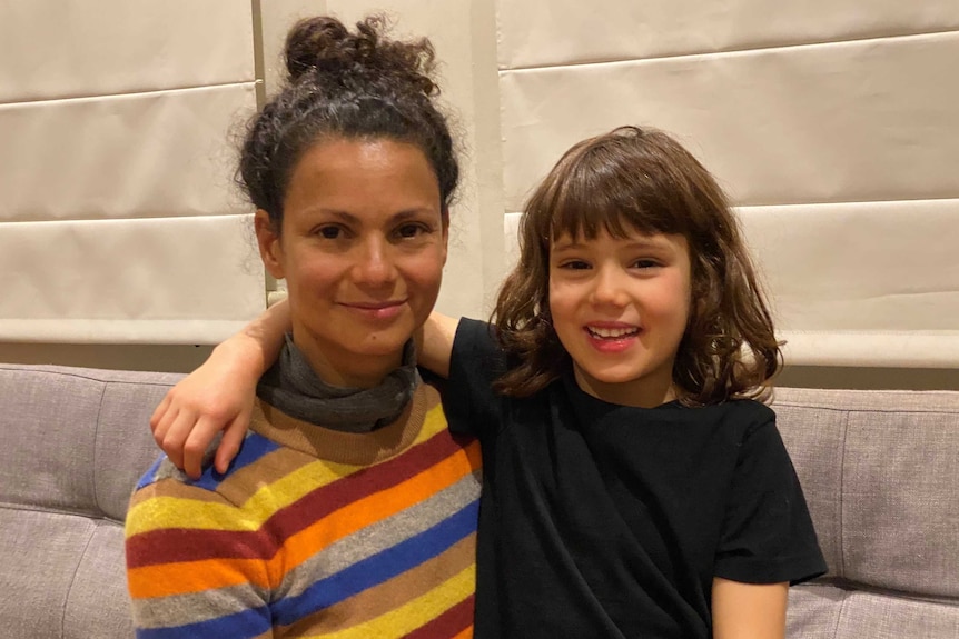 A young girl with dark hair sitting with her arm around her mother smiling.