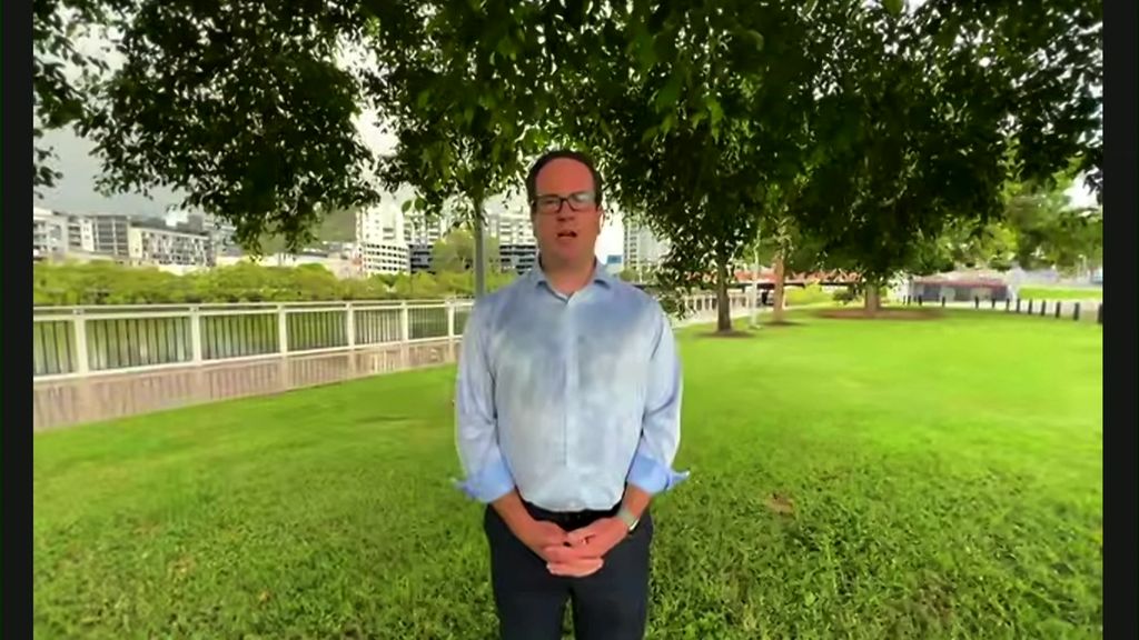 Man stands on lawn patch wearing blue button up shirt with hands softly clasped. 