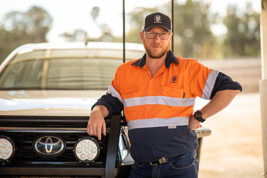 A man in a an orange high vis shirt and cap leans on a ute.