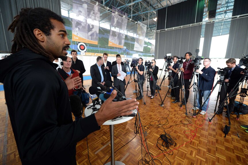 A football player stands in front of microphones and speaks to a media pack.