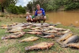 A man and two boys kneel on a riverbank in front of a veritable regatta of fish corpses.