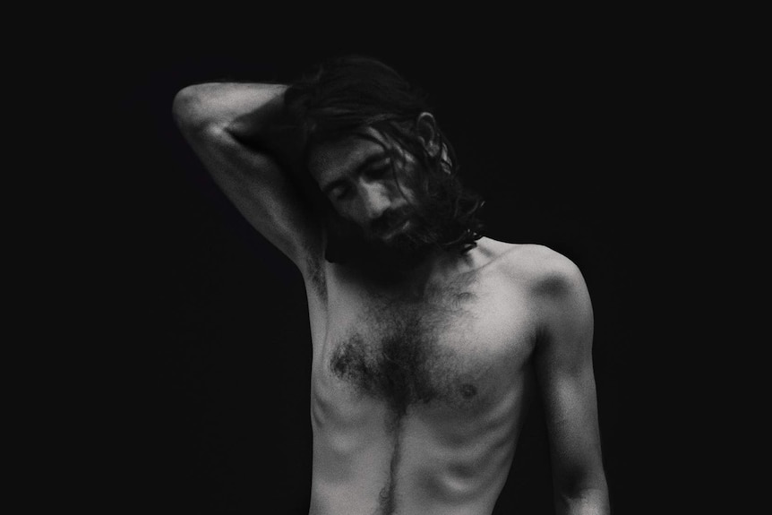 Black and white image with black background of Behrouz Boochani, shirtless and  looking downwards