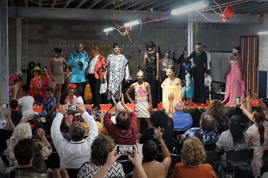 A group of models walk down the runway in front of a seated audience who are clapping and filming.