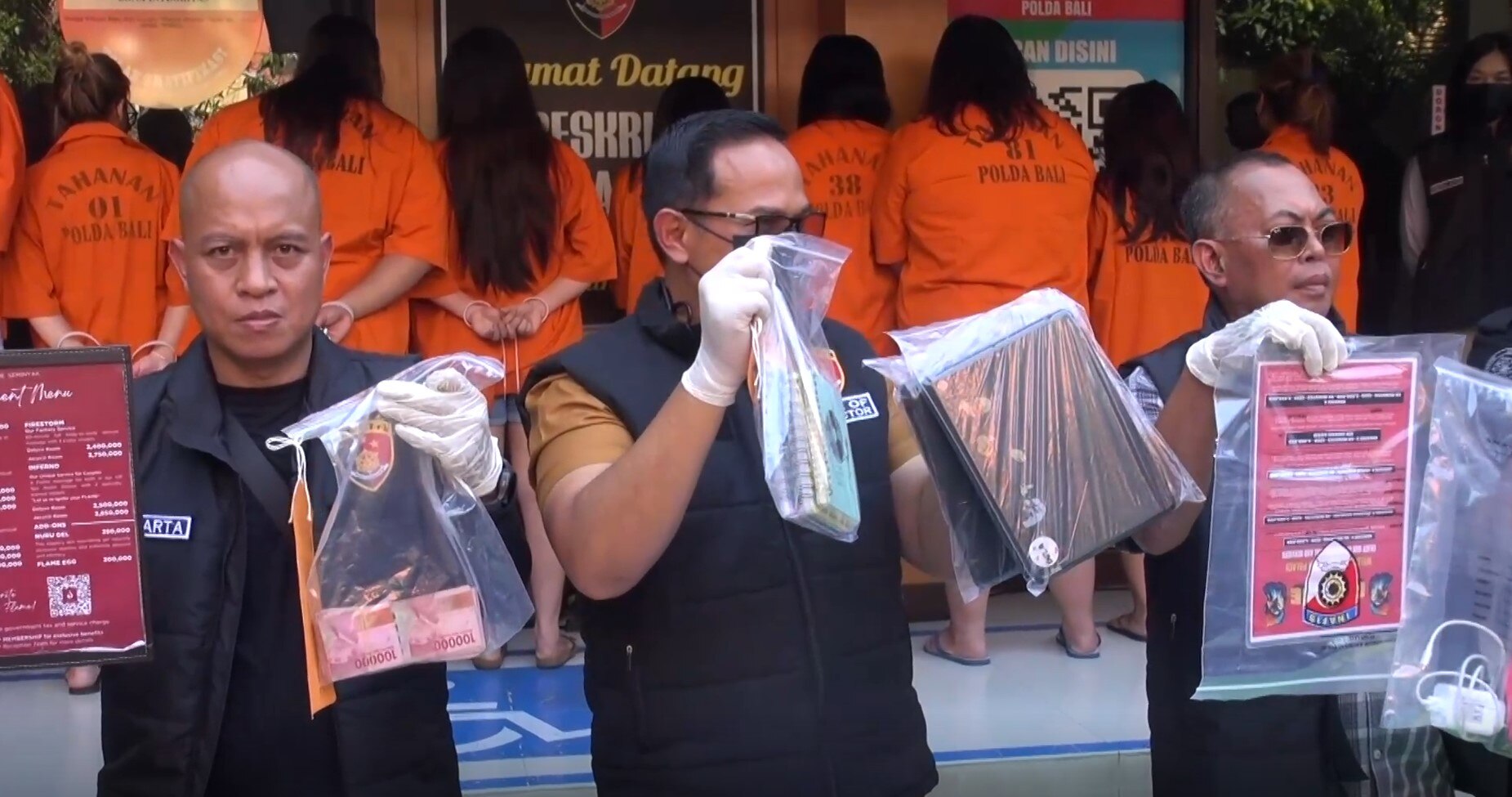 Three serious-looking police officers hold up plastic bags of evidence as people in orange shirts face a wall behind them.