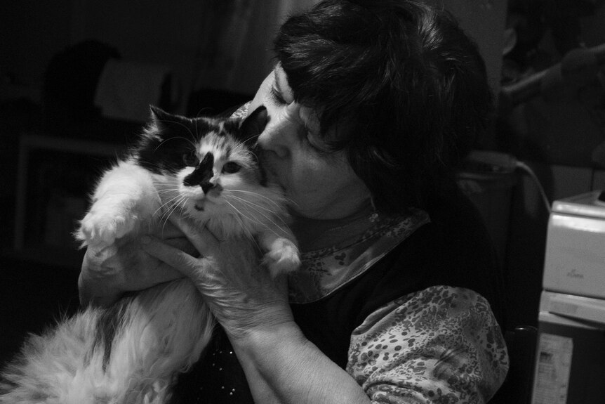 A black and white photo of a woman sitting at a table and holding up a fluffy cat, whose head she is kissing