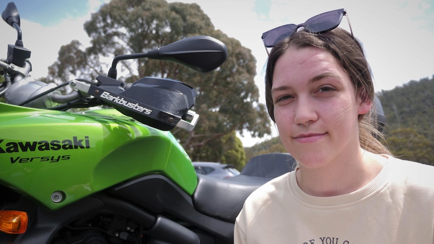 A young woman crouched in front of a green motorbike, smiling to camera.
