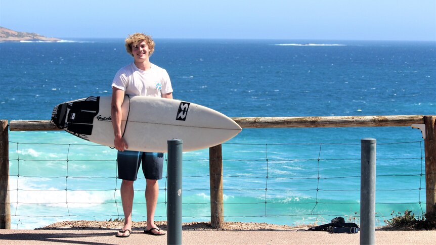 A smiling young man with curly blond hair, white t-shirt and blue shorts, stands near wooden railings in front of beach.