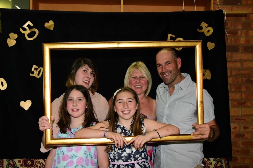 A family of five stand holding a picture frame smiling at the camera