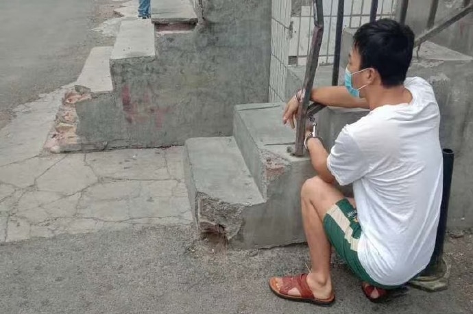 A person squats in place after being handcuffed to a rail beside a building in Urumqi.