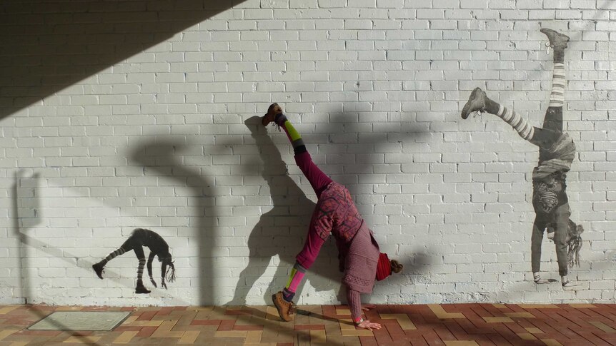 A woman does a handstand in between two images of herself doing a handstand pasted on a wall.