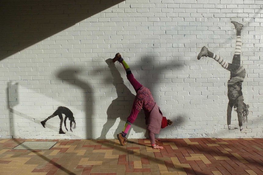 A woman does a handstand in between two images of herself doing a handstand pasted on a wall.