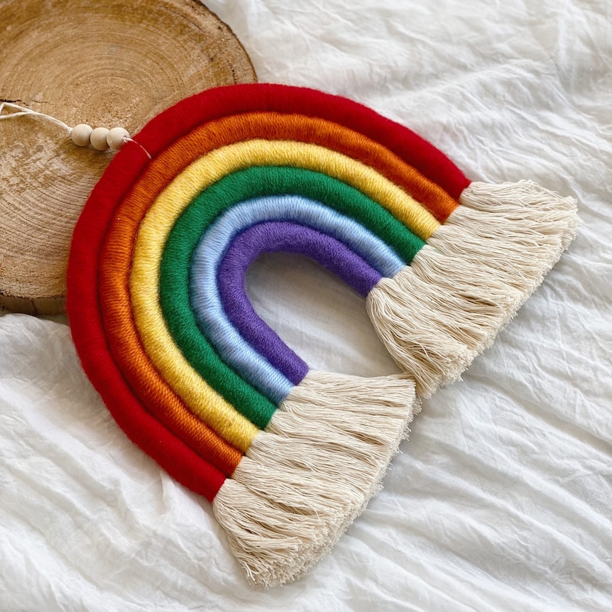 A tasseled macrame rainbow lies on a white cotton sheet next to a circular thin of wood. 