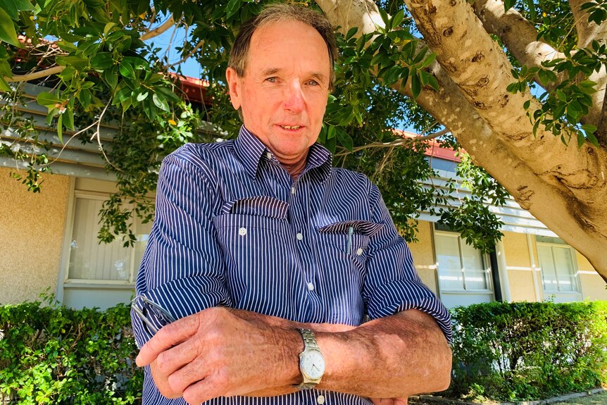 An older man in a checked shirt stands under a tree with his arms crossed.