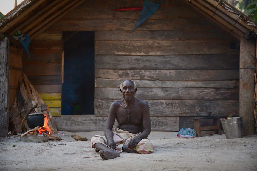 A man sits in the sand smiling, in background a pot hangs over a fire in front of a wooden house.