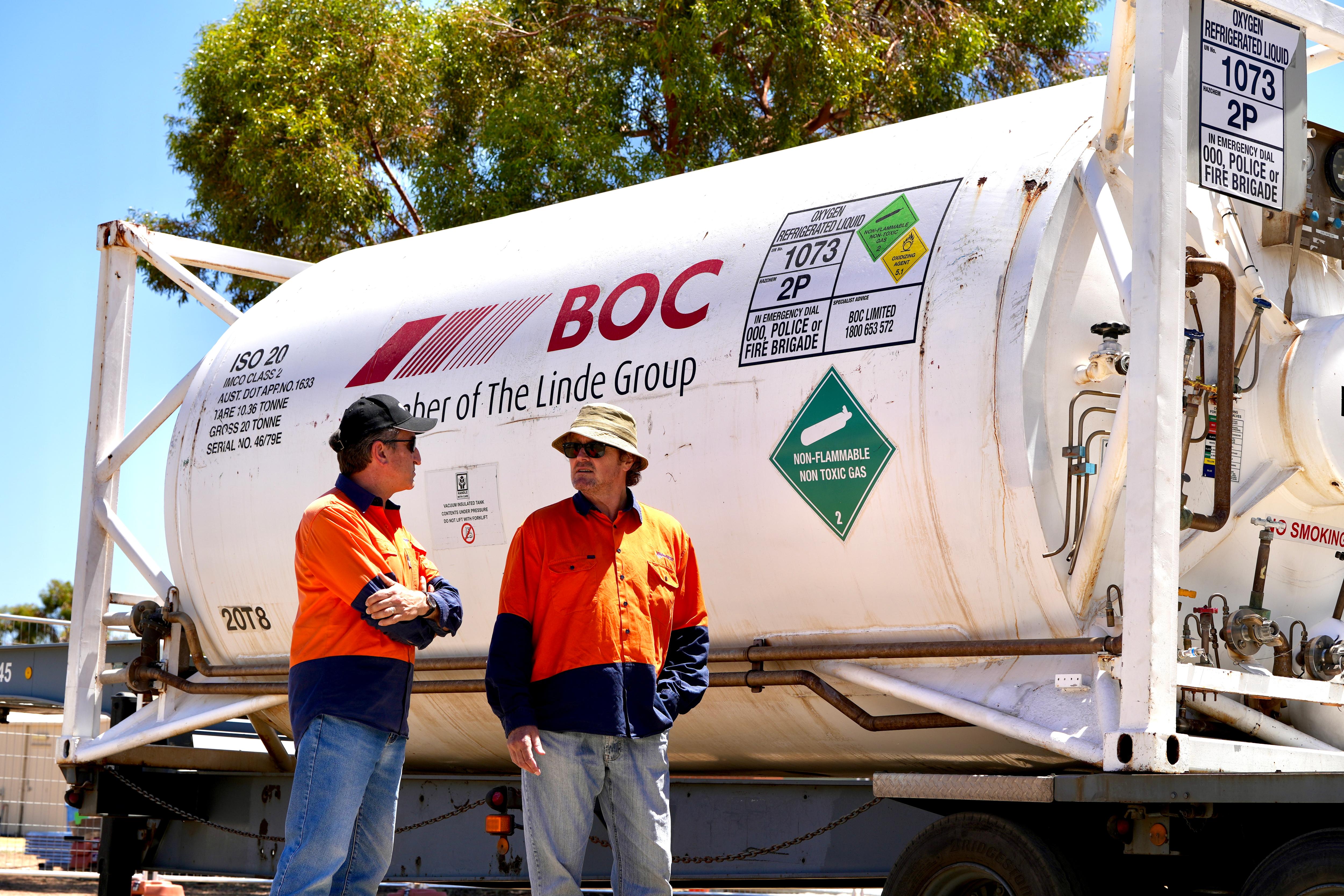 Two white men wearing hats, sunglasses and orange shirts stand in front of large white oxygen tank. 