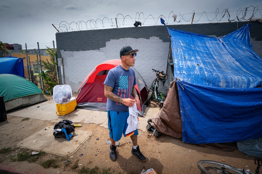 A man dressed in T-shirt and shorts stands next to a tent in an urban area