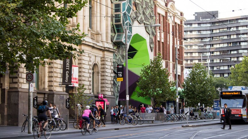 A colourful, geometric building sits between two older ones. People, bikes and trams going past.