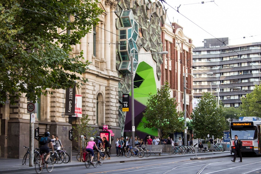 A colourful, geometric building sits between two older ones. People, bikes and trams going past.