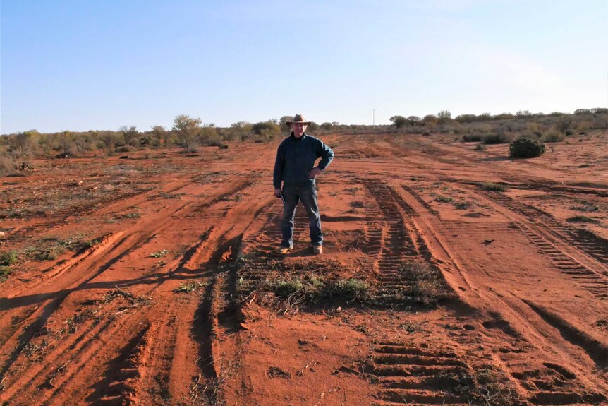 A man in rugged clothing and a hat stands in the red sand of the Australian outback.
