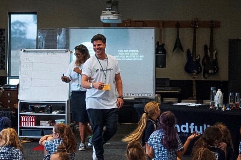 Matthew Runnalls smiles as he looks to the class in front of him. A woman stands behind him in a matching white shirt.