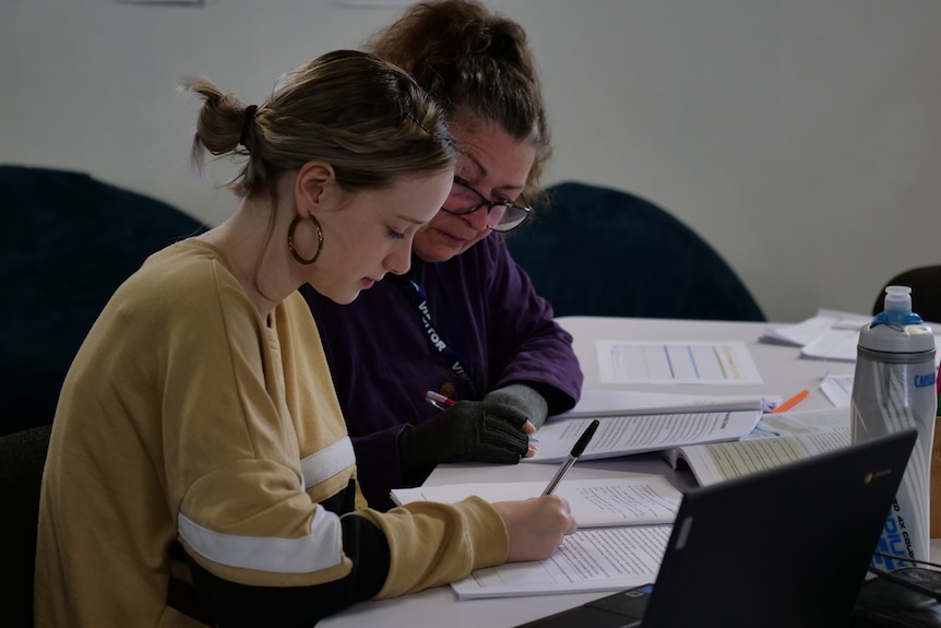 A female teacher sits with a teenage student at a desk, writing in books.