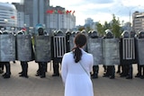 A woman in a white dress stands confronting a row of police with riot shields