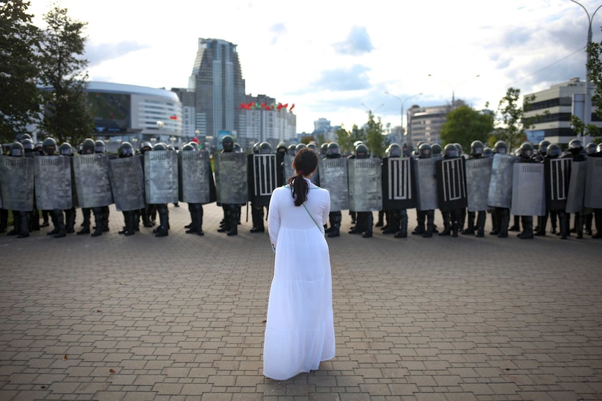 A woman in a white dress stands confronting a row of police with riot shields