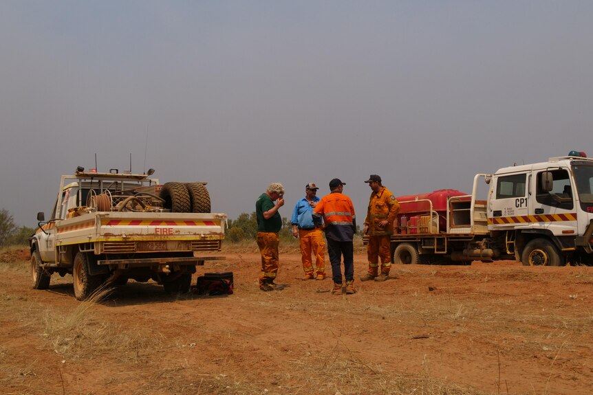 A small group of people in high vis standing on a dirt road between two parked vehicles and talking, with bushfire smoke ove